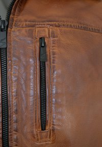 Braune Lederjacke mit strukturierter Oberfläche, ausgestattet mit einer schwarzen Reißverschlusstasche und kontrastierenden Nähten entlang der Nähte.