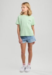 T-shirt verde chiaro a maniche corte con testo "SUMMER ESCAPE", abbinato a pantaloni corti in denim blu e sneakers bianche con calze a strisce rosse.