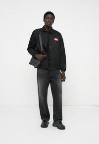 Svart knapp-up jakke med rød "DIESEL" logo, grå denim bukser og svarte joggesko, kombinert med en liten svart crossover-veske.