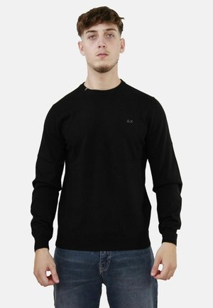 Maglione nero a maglia con maniche lunghe, scollatura a girocollo e un piccolo logo sul petto. Indossato con jeans blu, che mostrano un design semplice e aderente.
