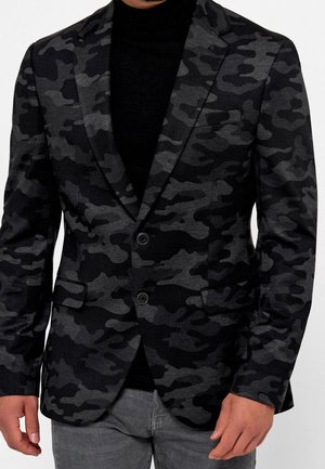 Mann trägt einen Blazer mit schwarzem und dunkelgrauem Camouflage-Muster über einem schwarzen Rollkragenpullover und grauen Jeans, steht mit entspannten Händen.