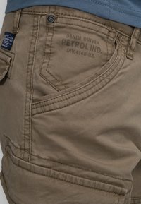 Khaki shorts met een gladde textuur, met een zijzak en een logo-label. De stof heeft een subtiele glans en versterkte stiksels.