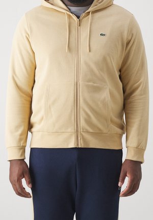 Mann trägt einen beigen Kapuzenpullover mit Reißverschluss und grünem Krokodil-Logo sowie dunkelblaue Hose, steht mit locker an den Seiten hängenden Armen.