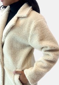 Abrigo de felpa esponjosa de color crema, con cuello notched, bolsillos frontales y tela texturizada, que cuenta con mangas largas y un ajuste relajado.