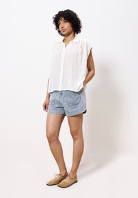 Weißes, kurzärmliges, kragenloses, knöpfbares Hemd; hellblaue Denim-Shorts; hellbraune Wildlederschuhe mit Schnürsenkeln; Model steht vor einem schlichten Hintergrund.