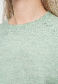 Pull vert clair à col rond côtelé. La texture semble douce et tricotée, avec un motif chiné subtil.