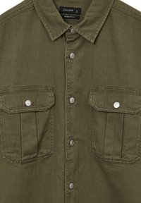 Camicia di jeans verde oliva con chiusura a bottoni, due tasche sul petto e bottoni a pressione argentati, caratterizzata da un design a vestibilità regolare e tessuto testurizzato.