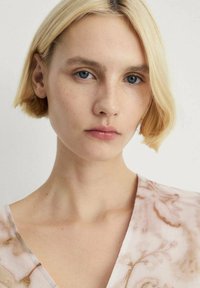 Gros plan sur une personne ayant les cheveux blonds courts, des yeux bleus et de légères taches de rousseur, portant un haut en col en V avec un motif floral rose et une texture douce.