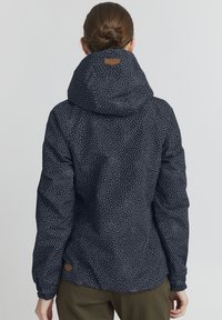 Giacca blu navy con pois bianchi, dotata di un ampio cappuccio e dettagli in pelle marrone. Il materiale appare tessurizzato e isolato.