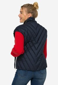 Marinblå quiltad pufferväst med hög krage, dragkedja och chevronmönster, tillsammans med en röd stickad tröja och blå jeans.