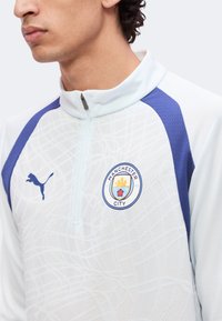 Άνδρας που φοράει αθλητικό τοπ της Manchester City σε λευκό και μπλε με μισό φερμουάρ και λογότυπο της Puma στο στήθος.