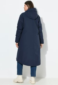 Námořnický modrý dlouhý kabát s kapucí, s hladkým, voděodolným povrchem. Nosený s světle barevnými robustními botami a modrými džíny.