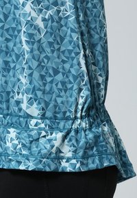 Haut de sport bleu avec un motif géométrique aux nuances variées de bleu. Il possède un ourlet froncé et un tissu lisse et extensible.