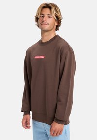 Bruine sweatshirt met een ronde hals en lange mouwen, met een rood Quiksilver logo op de borst. Zachte stof, ontspannen pasvorm.
