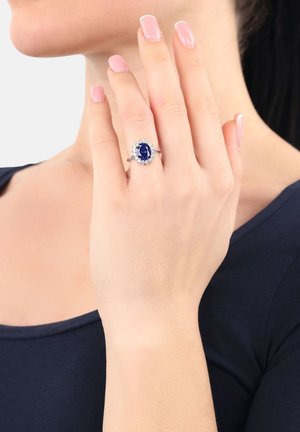 Ovaler blauer Edelsteinring, umgeben von kleineren klaren Steinen, in einem Silberring eingefasst. Hand in pose vor neutralem Hintergrund.
