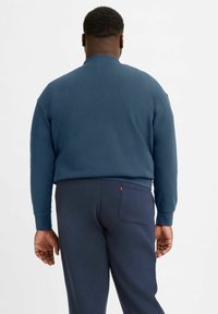 Felpa blu con collo a giro e polsini a costine, abbinata a pantaloni scuri con tasca posteriore e dettaglio del logo. Tessuto morbido.