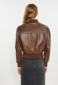 Jachetă bomber din piele maro, cu guler și manșete ribbate. Prezintă un tiv strâns și o textură elegantă cu un finisaj neted.