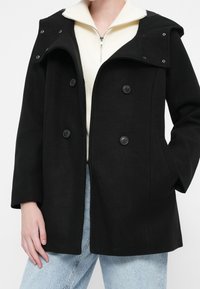 Woman wearing a black double-breasted coat over a white zip-up sweater and light blue jeans, with one hand in pocket.