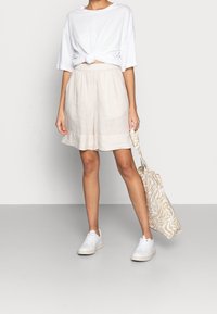 T-shirt bianco legato, pantaloni corti beige in lino e sneakers bianche. Tiene una borsa tote beige con un sottile motivo a zebra. Texture liscia, vestibilità casual.