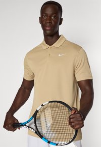 Világosbarna, rövid ujjú póló, két gombbal és fehér Nike logóval. A modellen teniszütő van, kék és fekete markolattal.