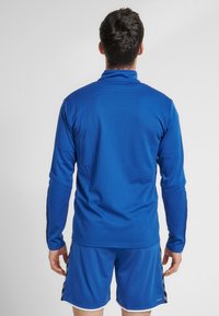 Maglietta atletica blu a maniche lunghe con finitura testurizzata, colletto alto e accenti a strisce nere lungo le maniche, abbinata a pantaloni corti coordinati.