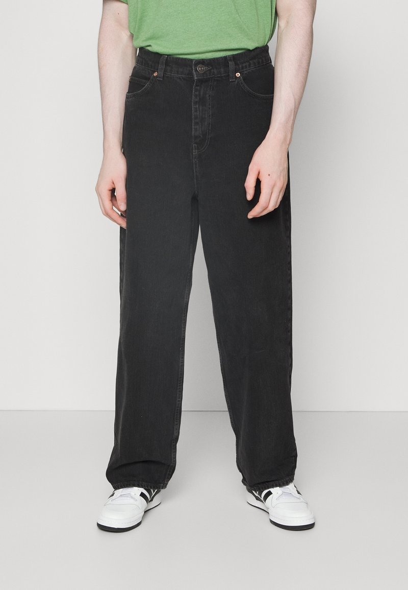 Černé džíny rovného střihu s knoflíkovým zapínáním, dvěma předními kapsami a jemným prošíváním na švech, doplněné bílými teniskami.