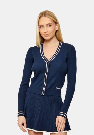 Donna che indossa un cardigan blu navy con bottoni, con strisce bianche e rosa, abbinato a una gonna plissettata coordinata su uno sfondo semplice.