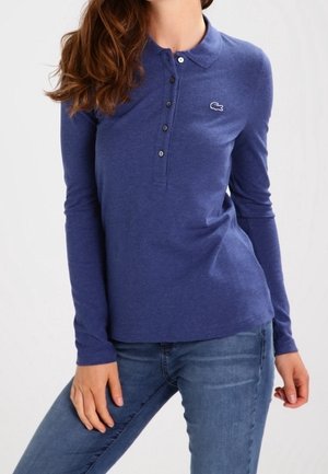 Femme portant un polo bleu ajusté à manches longues avec des boutons et un petit logo crocodile, assorti à un jean bleu. Haut du corps visible.