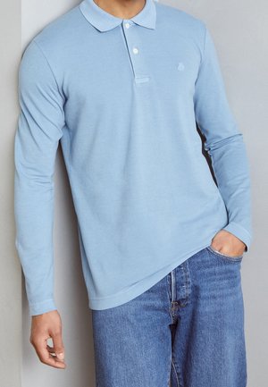 Mand iført en lyseblå langærmet polotrøje med hvide knapper og blå jeans, stående med den ene hånd i lommen.