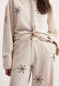 Beige skjorta och byxor i ett set, tillverkade av strukturerad tyg, med mörkbrun blommig broderi. Knappdetaljer på skjortan och byxorna.