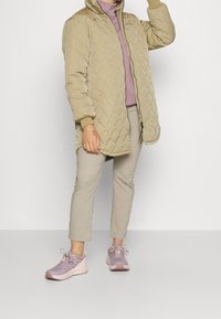 Beżowy pikowany długi płaszcz z wysokim kołnierzem, jasnofioletowa bluzka z długim rękawem, jasnobeżowe spodnie oraz różowawe buty sportowe.