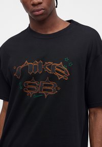 Νέο άτομο που φοράει μαύρο μπλουζάκι Nike SB με πορτοκαλί περίγραμμα κειμένου και μικρά πράσινα αστέρια μπροστά.
