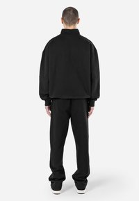 Felpa nera oversize con colletto alto, abbinata a pantaloni larghi in coordinato. Il tessuto appare morbido, con una superficie liscia e dettagli minimi.