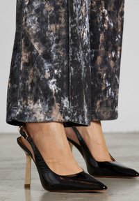 Schwarze Lackleder Slingback-High Heels mit spitzer Zehe, goldfarbener Stiletto-Ferse und strukturierten Oberflächen. Graue und braune gemusterte Hose sichtbar.