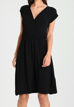 Nainen, joka pukeutunut polvipituiseen mustaan mekkoon, jossa on V-kaula-aukko, lyhyet hihat ja kerrostettu vyötärö, seisoo yksinkertaisen taustan edessä.