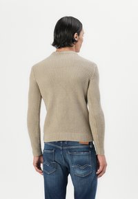 Pullover beige lavorato a maglia con un motivo testurizzato, collo a giro, e polsini a costine, abbinato a jeans blu con dettagli di cucitura sulle tasche posteriori.