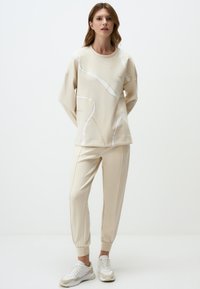 Beige sweatshirt met abstracte witte lijnen, gecombineerd met bijpassende beige joggingbroek. Zachte textuur, relaxte pasvorm en elastische manchetten. Witte sneakers.