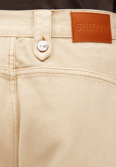 Κοντινό πλάνο σε μπεζ τζιν παντελόνια που δείχνει την θηλιά της ζώνης, ασημένιο κουμπί με το χαραγμένο "Stieglitz" και καφέ δερμάτινο έμπλαστρο με το όνομα της μάρκας.