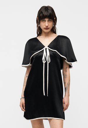 Femme aux cheveux noirs portant une robe en satin noire avec des bordures blanches, des manches capes courtes, et un détail noué à l'avant, debout devant un fond uni.