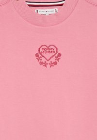 Ružová mikina má okrúhly výstrih, vyšívaný dizajn srdca s nápisom "TOMMY HILFIGER" a kvetinové akcenty v tmavšej ružovej.