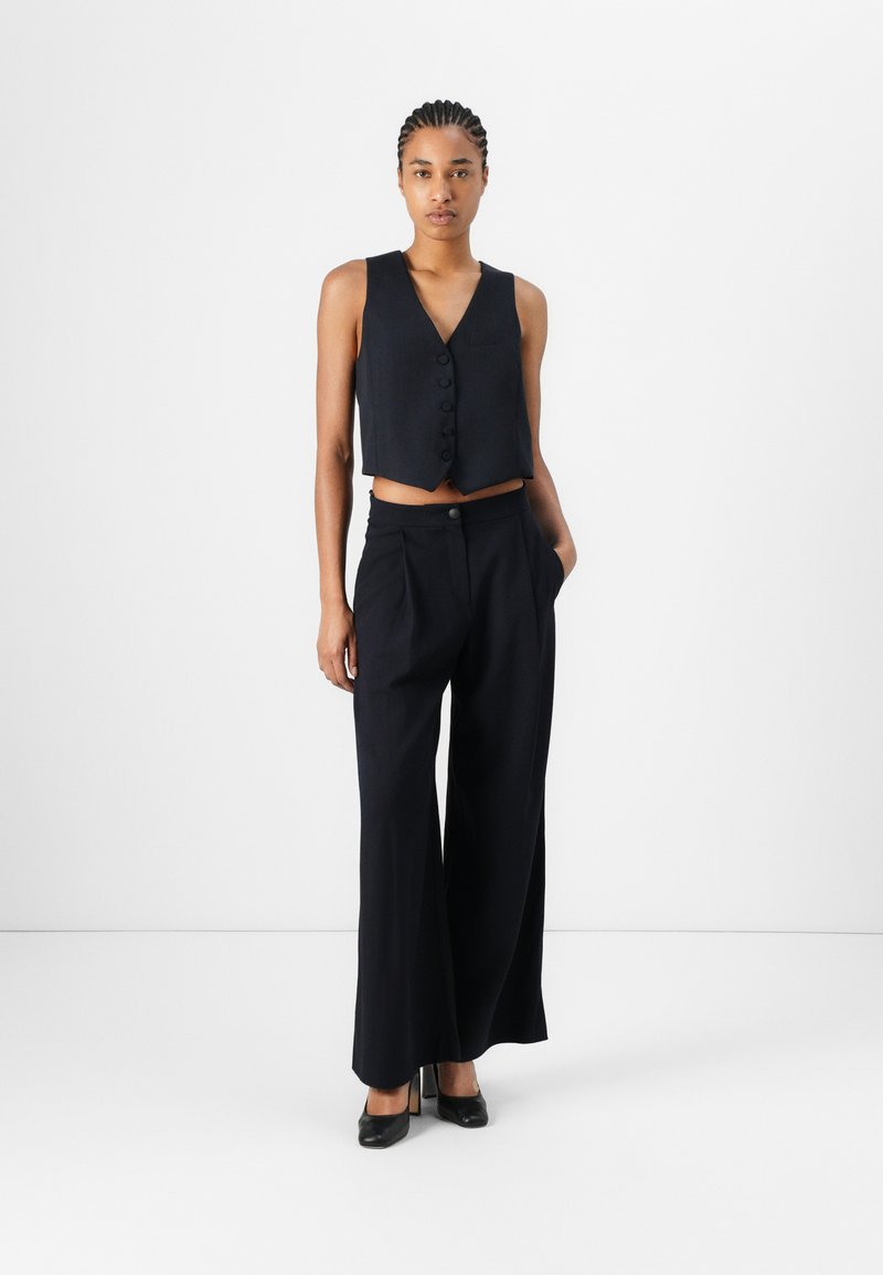 Zwart cropped vest met een v-halslijn en knoopdetails, gecombineerd met hoge taille, wijde broekspijpen. Gesloten zwarte hakken maken de look compleet.