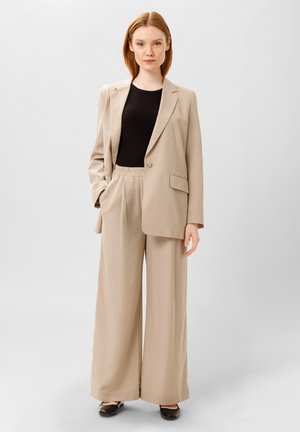 Beige op maat gemaakte blazer met één knoop en zijzakken, gecombineerd met beige wijde broek met een elastische tailleband. Zwart top eronder.