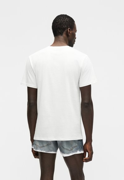 Άνδρας που στέκεται με πλάτη, φοράει απλό λευκό κοντομάνικο μπλουζάκι και ανοικτό γαλάζιο ξεφτισμένο τζιν σορτς, μπροστά σε απλό φόντο.