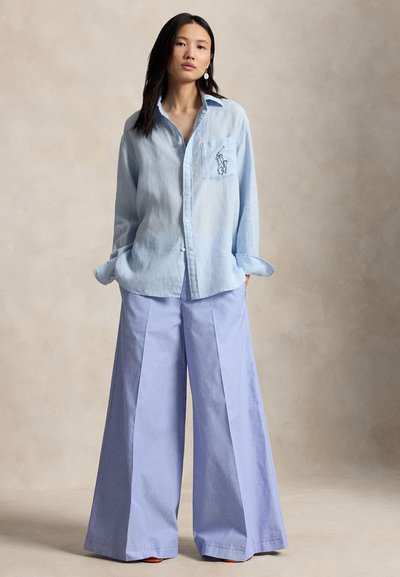 Žena stojaca s rukami v vreckách, oblečená v svetlomodrej voľnej košeli na gombíky a širokých modrých nohaviciach na jednoduchom pozadí.