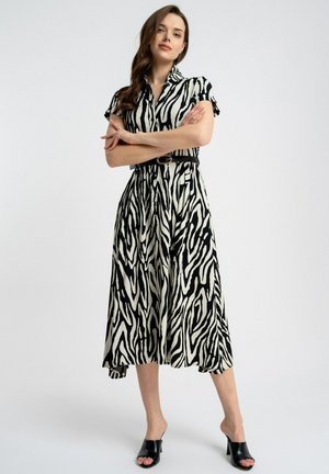Kobieta stojąca z założonymi na piersiach rękami, ubrana w midi sukienkę w biało-czarne paski zebry, z czarnym paskiem i czarnymi sandałami na obcasie na białym tle.