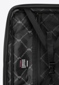 Intérieur d'un sac de voyage noir présentant une texture lisse, des poches en mesh et une étiquette de marque avec un motif de logo répétitif. Compartiments zippés inclus.