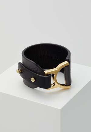 Schwarzes Lederarmband mit breitem Band und goldfarbenem Metallschnallen-Detail, präsentiert auf einem weißen Sockel vor einem schlichten grauen Hintergrund.