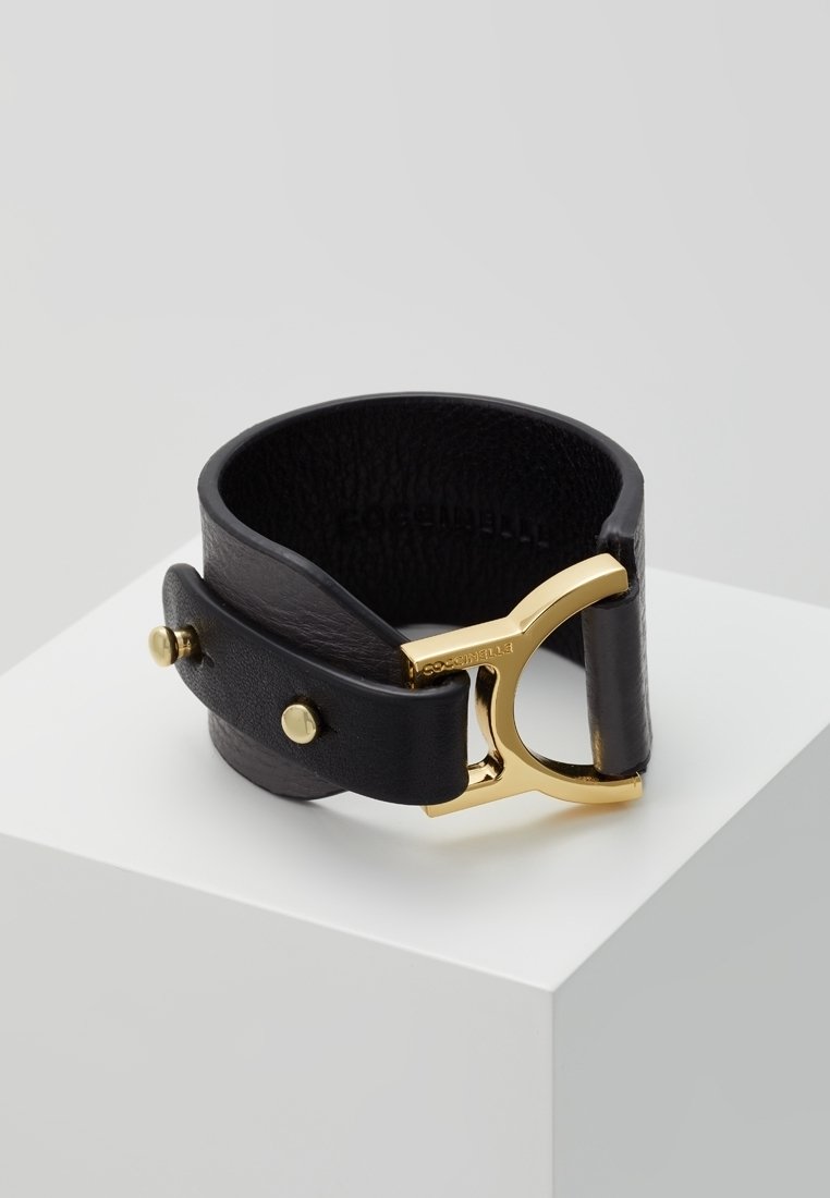 Bracelet en cuir noir avec large bande et boucle en métal doré, présenté sur un socle blanc sur fond gris uni.