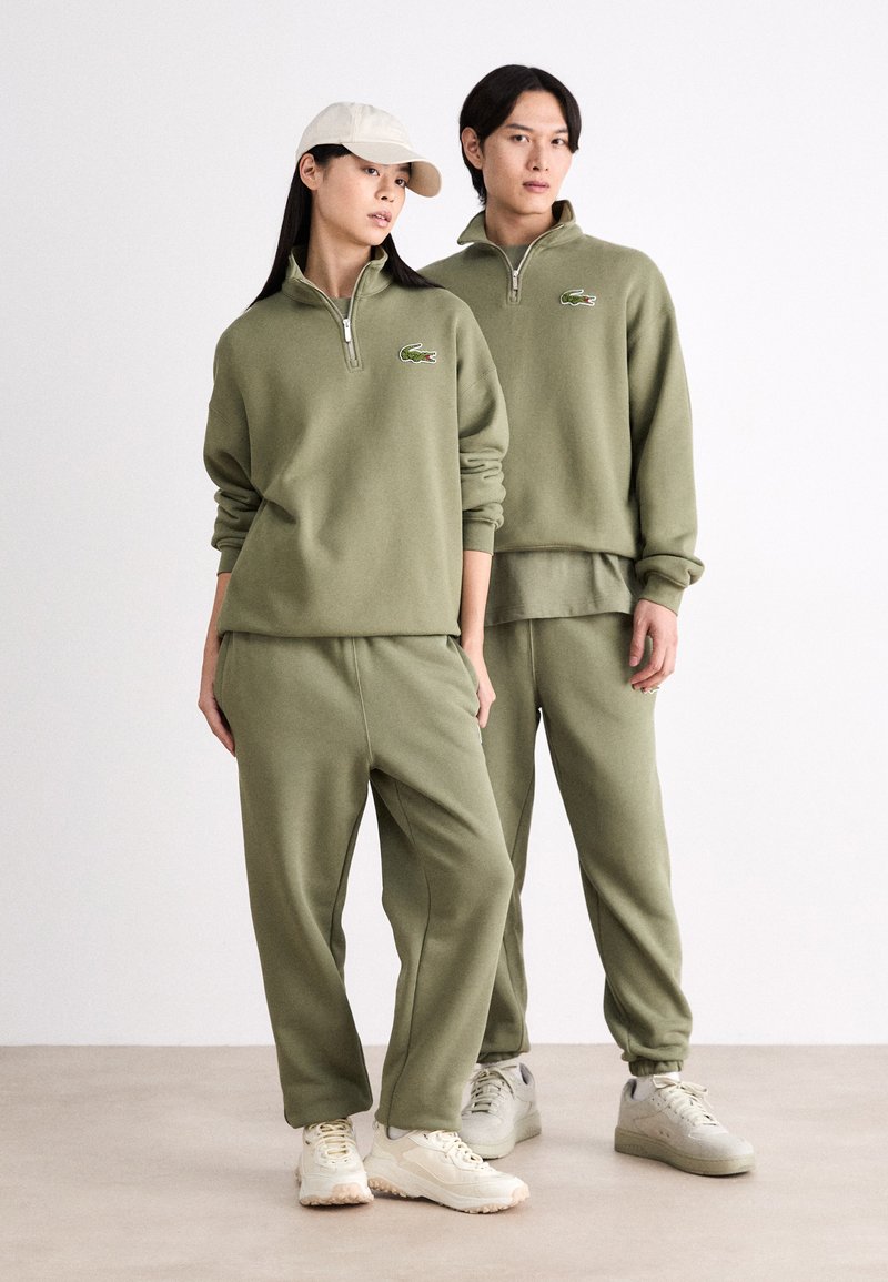 Sweatshirts zippés en coton vert olive avec un pantalon de survêtement assorti. Présente un logo brodé et une coupe décontractée, accompagnée de baskets de couleur neutre.