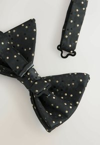Black bow tie with textured fabric, featuring gold and black polka dots. Adjustable black strap with a metal clasp.